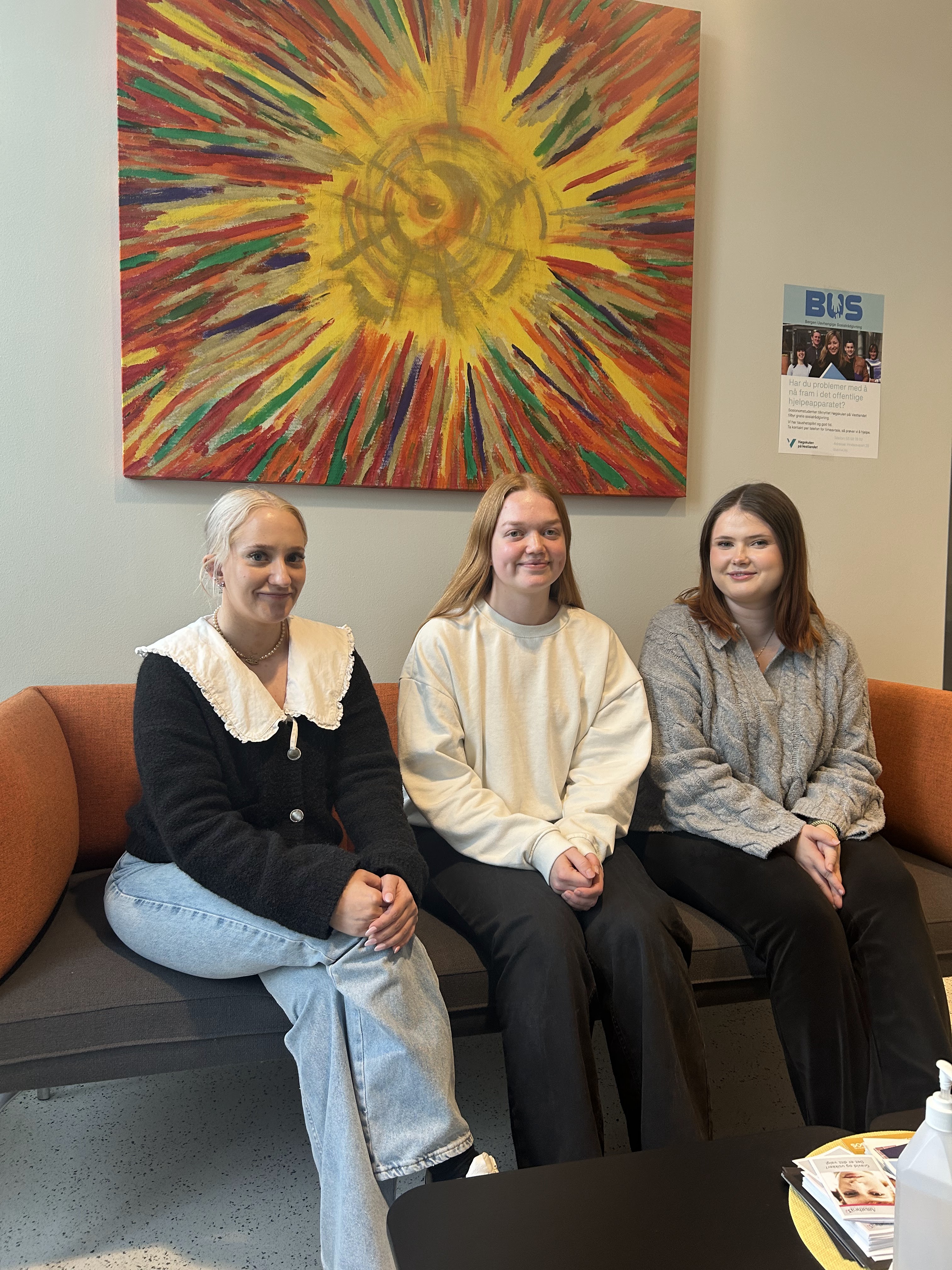 Tre unge mennesker i en sofa, og et stort maleri på veggen bak dem.