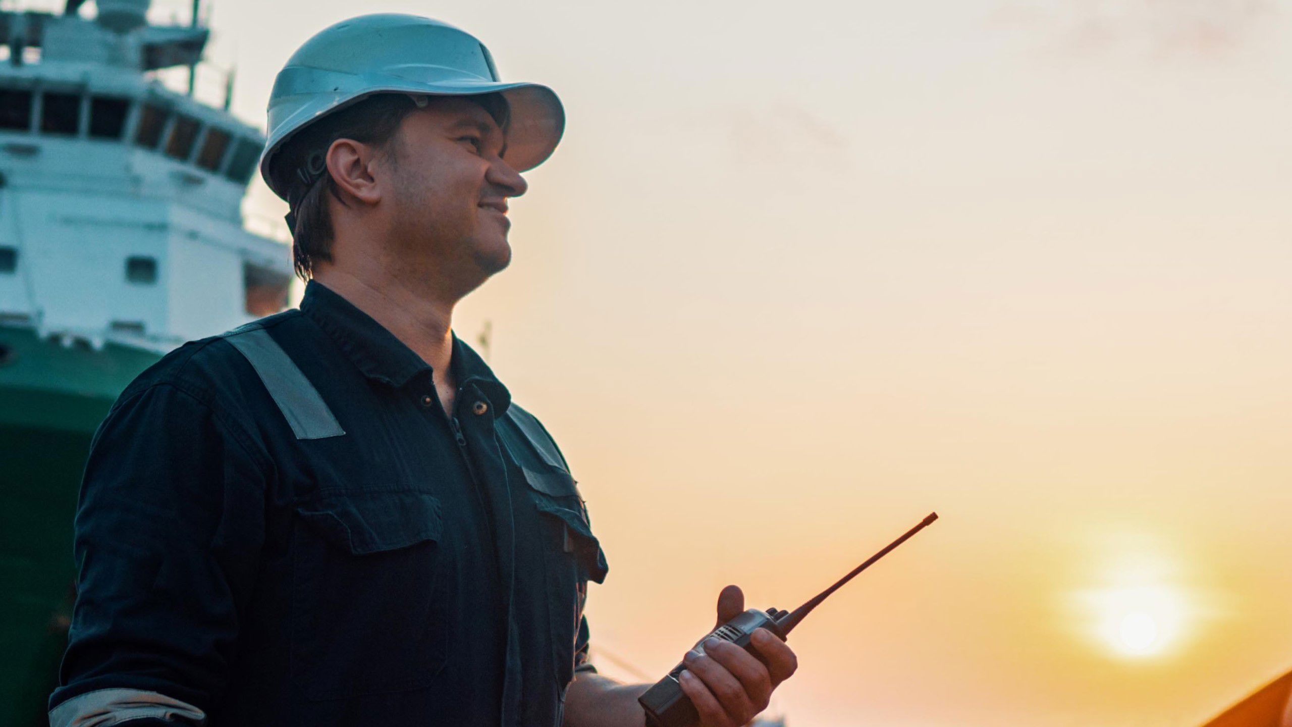 Mann i arbeidsklær med kommunikasjonsradio i hånda med skip i bakgrunnen.