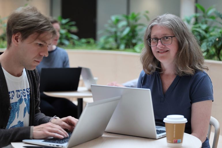 En kvinne og en mann sitter med hver sin pc rundt et bord.