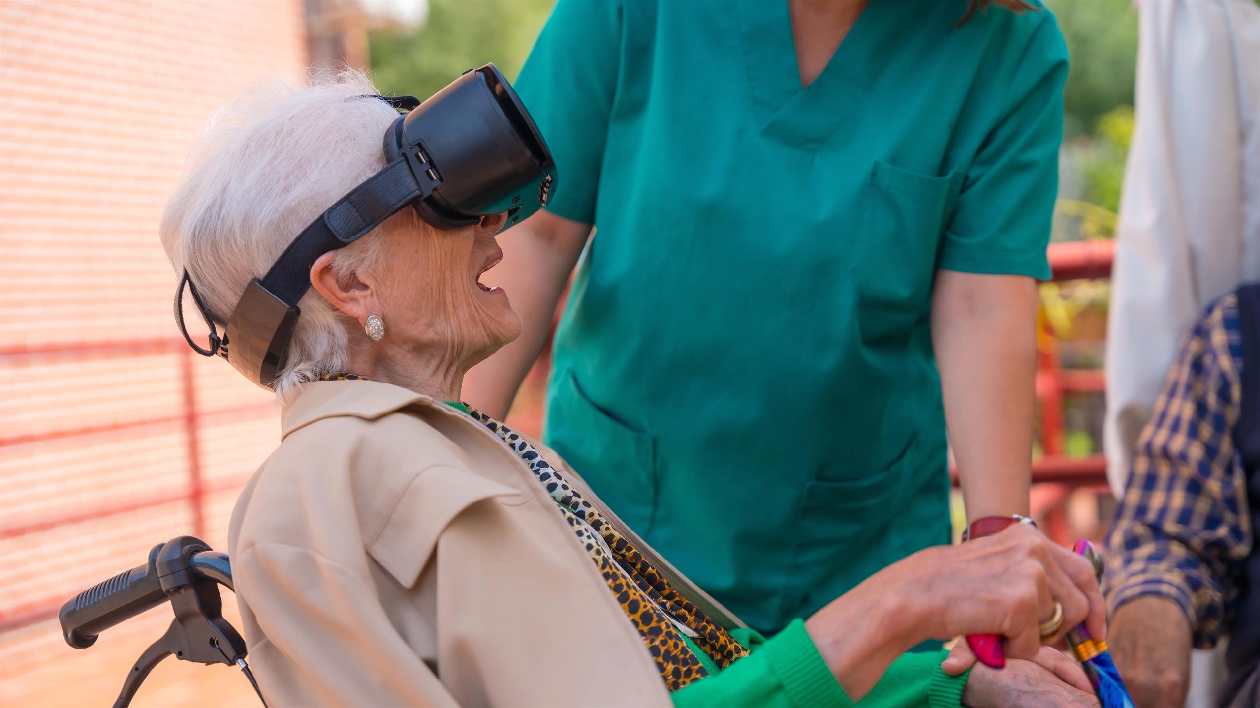 Eldre kvinne sitter i rullestol med VR-briller på.  Helsearbeider i grønn uniform i bakgrunnen. 