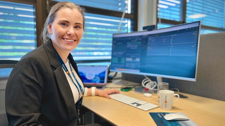 Smilande kvinne ser i kamera. Ho sit på kontoret og jobbar på PC'en.