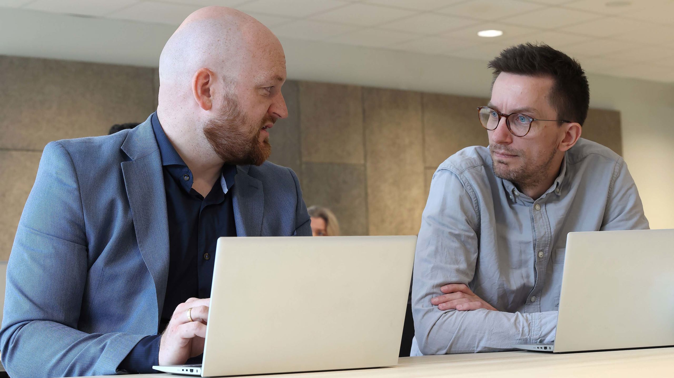 To menn sitter vet et bord med bærbare pcer foran seg. De ser på hverandre.