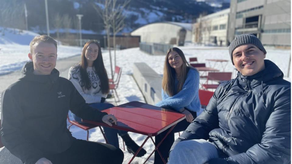 Fire fornøyde studenter sitter rundt et kafebord ute i sola, og smiler til kameraet. 