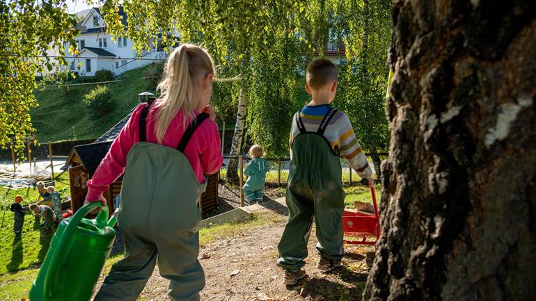 Barn i barnehage bærer vannkanne og triller trillebår.