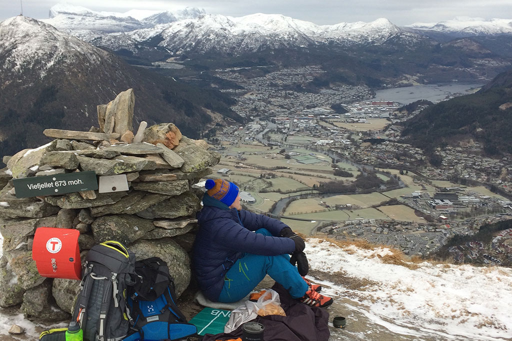 Mann sittande på fjelltopp med raud turpostkasse bak.
