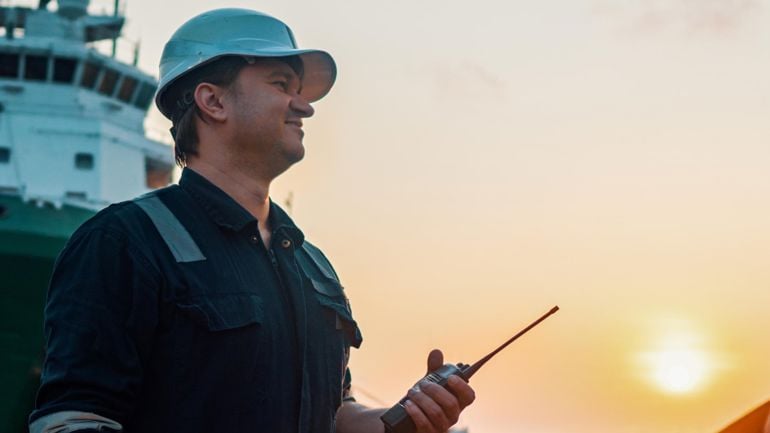 Mann i arbeidsklær med kommunikasjonsradio i hånda med skip i bakgrunnen.